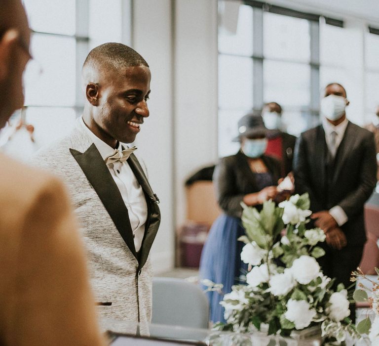 Smiling groom in grey Moss Bros suit with white shirt and satin bow tie stands at the altar with bride