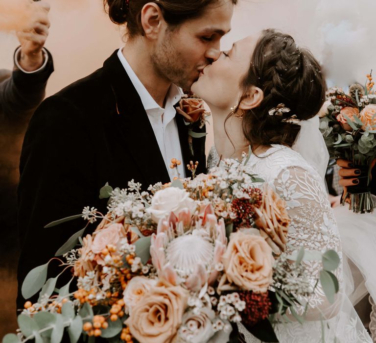 Bride and groom kissing on wedding day with orange smoke canon in the background and peach rose wedding bouquet 