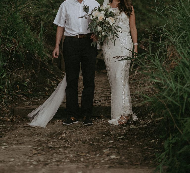 Bride walks with her father on wedding day through greenery 