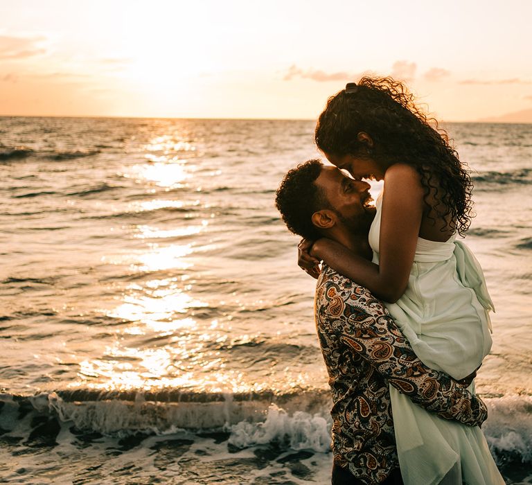 Groom lifts bride as she leans down to kiss him on the beach