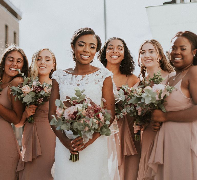 Bride in a lace wedding dress standing with her bridesmaids in blush pink dresses holding their red and pink bouquets 