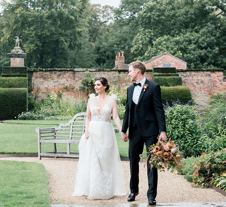 Groom in a black-tie suit holding hands with his bride in a tulle and lace romantic wedding dress walking through the gardens at Dorfold Hall