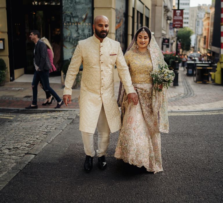 Bride in a pastel Bengali wedding dress crossing the road in Birmingham with her husband in a light gold coat 