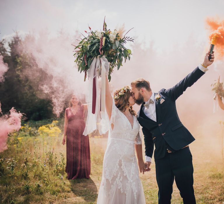 Bride & groom stand with wedding party whilst holding colourful smoke bombs