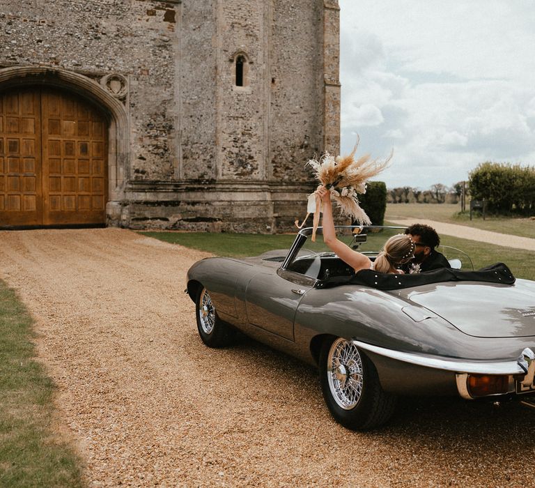 Bride and groom kissing in a vintage wedding car with the bride holding her dried flower bouquet in the air 