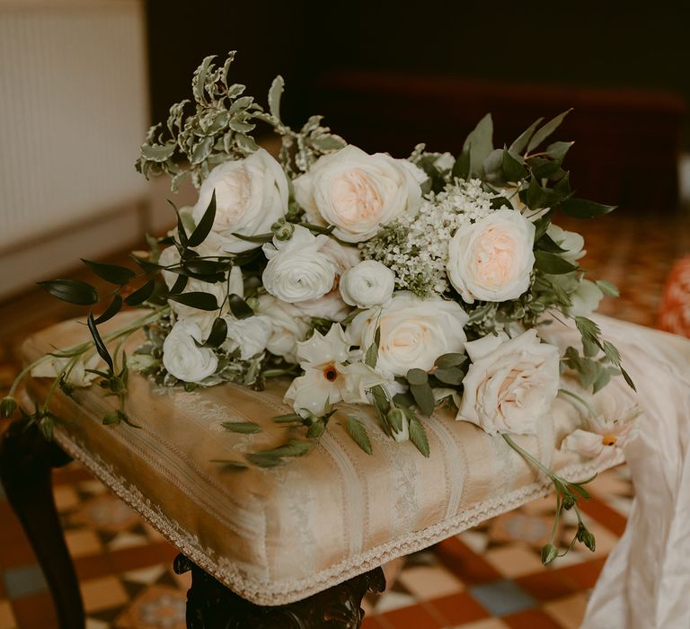 Bridal bouquet filled with white roses and green foliage in classic styling lays upon chair 