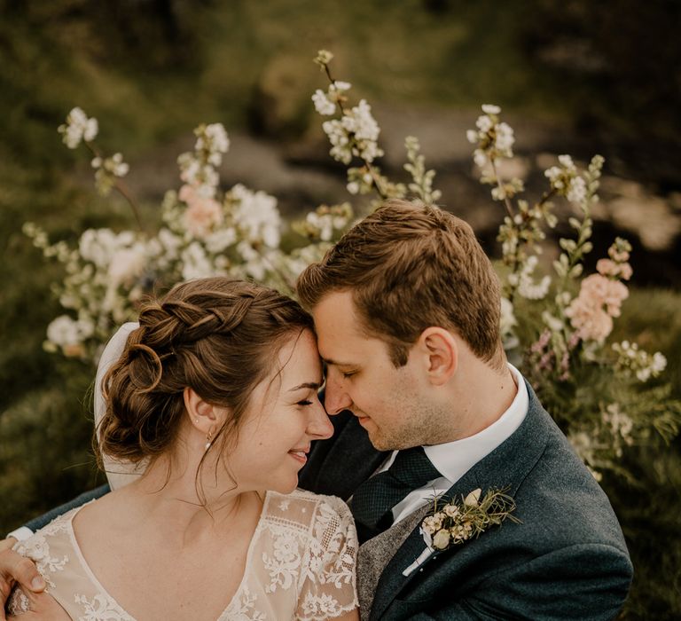 Bride in lace top wedding dress with capped sleeves and satin skirt rests forehead on groom in navy suits head whilst they sit down on clifftop after Dunluce Castle wedding