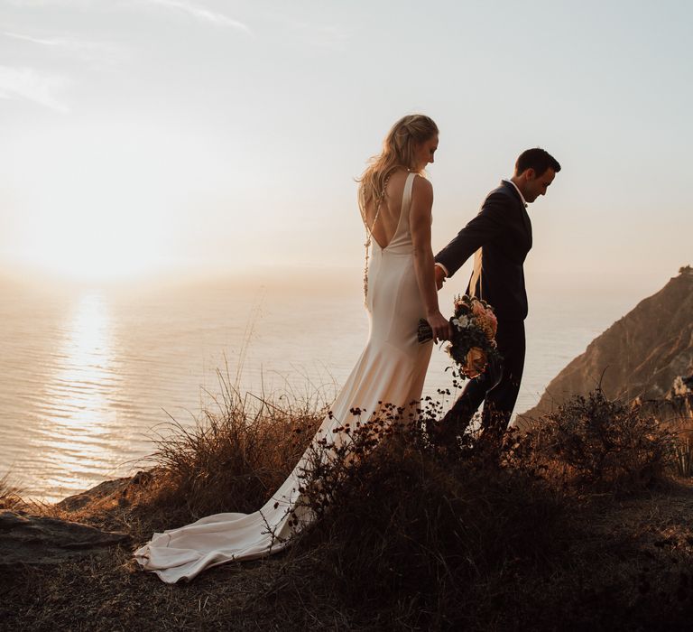 Bride & groom walk along the cliffs as the sun sets in the background across the ocean