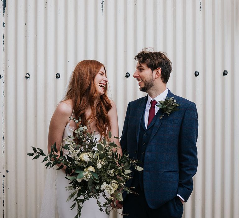 Bearded groom in a navy with red check wool suit looking at his smiley bride in a satin slip Grace Loves Lace wedding dress as she holds a green and white bouquet 