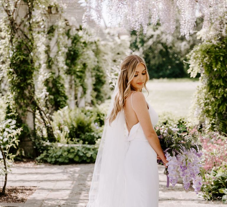 Bride stands beneath pergola with hanging wisteria and lilac florals above her whilst wearing Ava Mia Couture gown