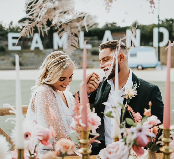 Groom in a open collar white shirt and black suit kissing his brides hand in a sparkly wedding dress at their outdoor tablescape at Dreamland Margate 