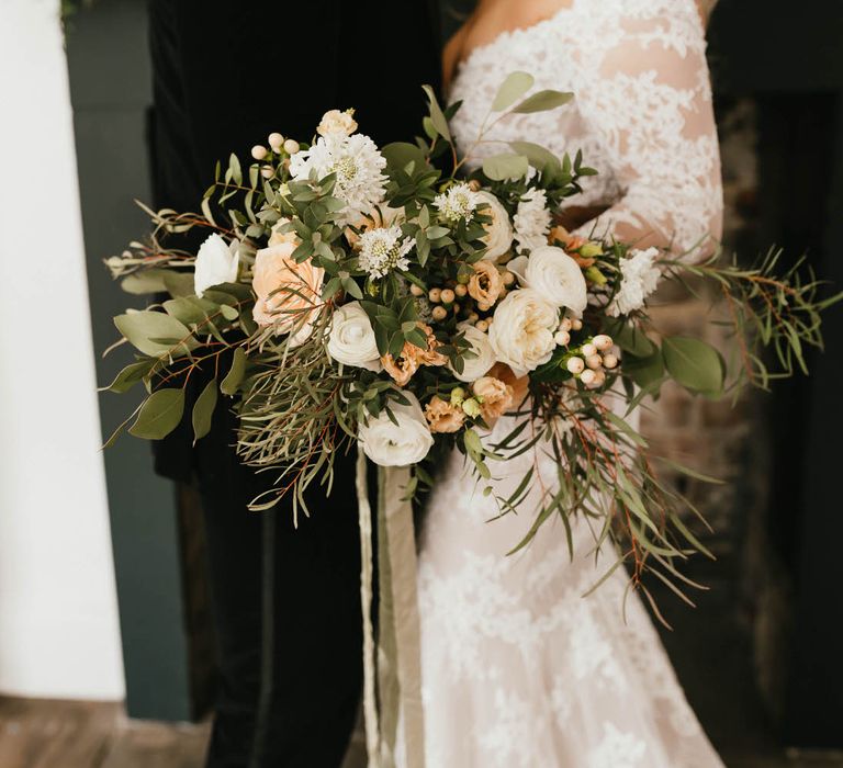 Bride holding a large bouquet of peach and white roses with foliage and wild grasses