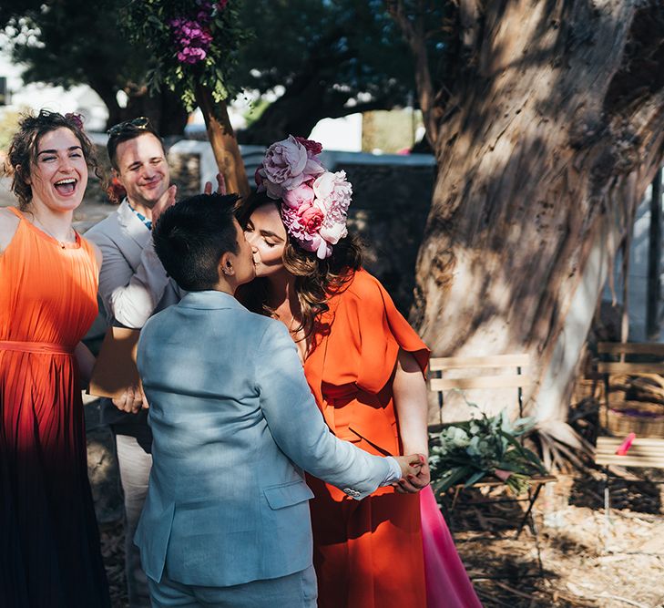 Brides kiss outdoors on their wedding day as wedding guests celebrate beside them