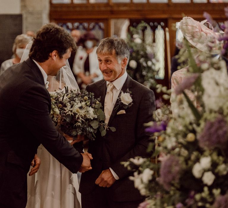 Groom in dark brown suit shakes hand of smiling father of the bride at the church altar 