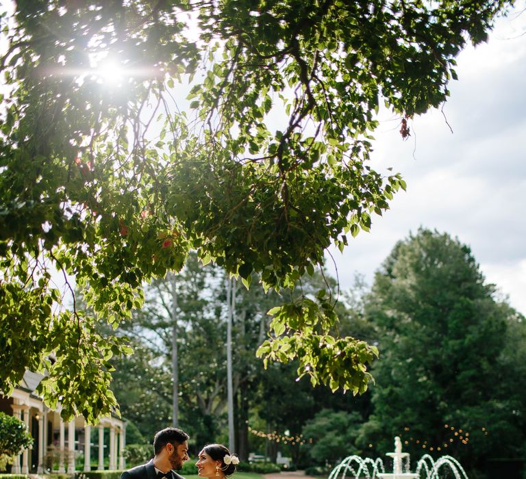 Bride & groom stand outdoors as the sun shines through the trees on their wedding day