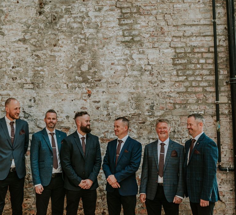 Groom stands with his groomsmen in front of industrial style brick wall whilst all wearing different suits in different tones of blue