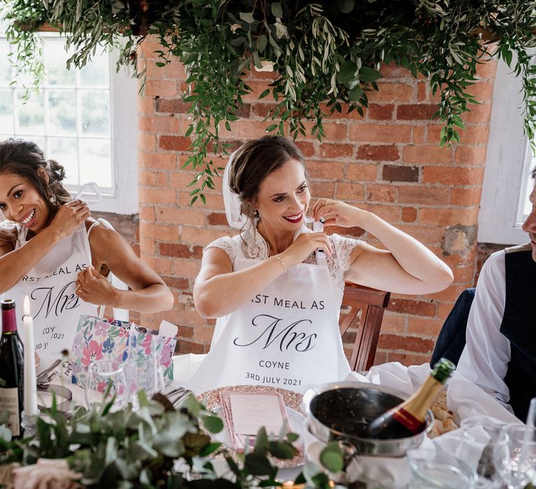 Two brides sat at top table both put on personalised white aprons reading 'First meal as Mrs Coyne 3rd July 2021' in red brick reception room at The West Mill Derby