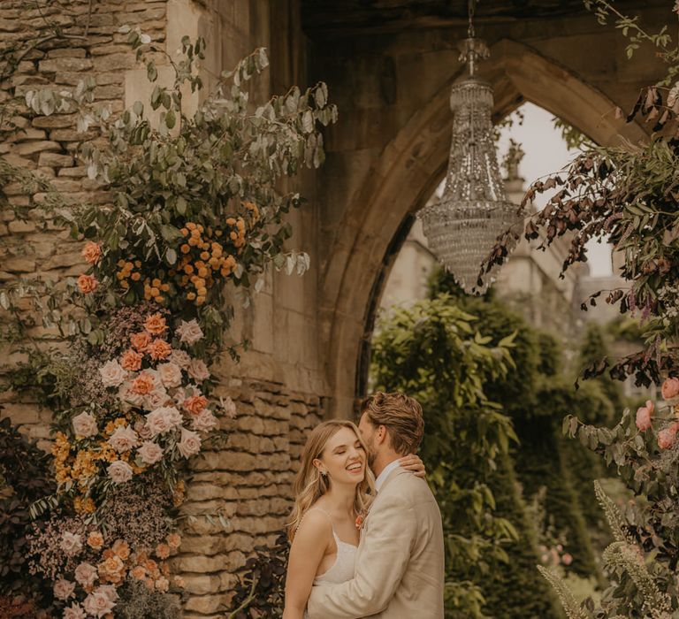 Groom in a beige suit embracing his bride in an embellished wedding dress by a pastel flower arrangement decorating the castle arch at Euridge Manor 