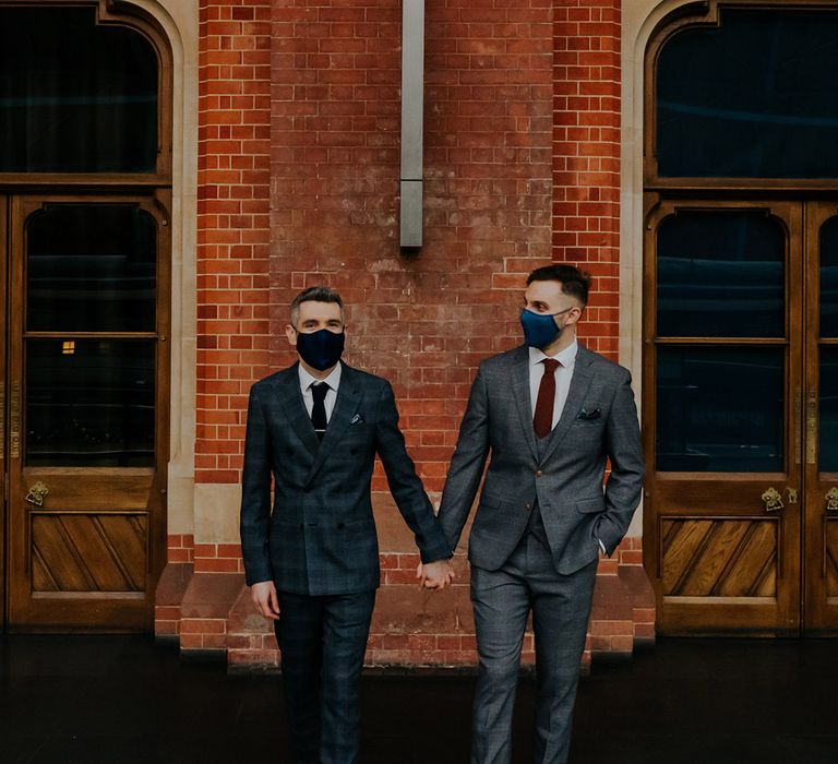 Grooms stand in front of brick wall whilst wearing face coverings in stylish suits on their wedding day