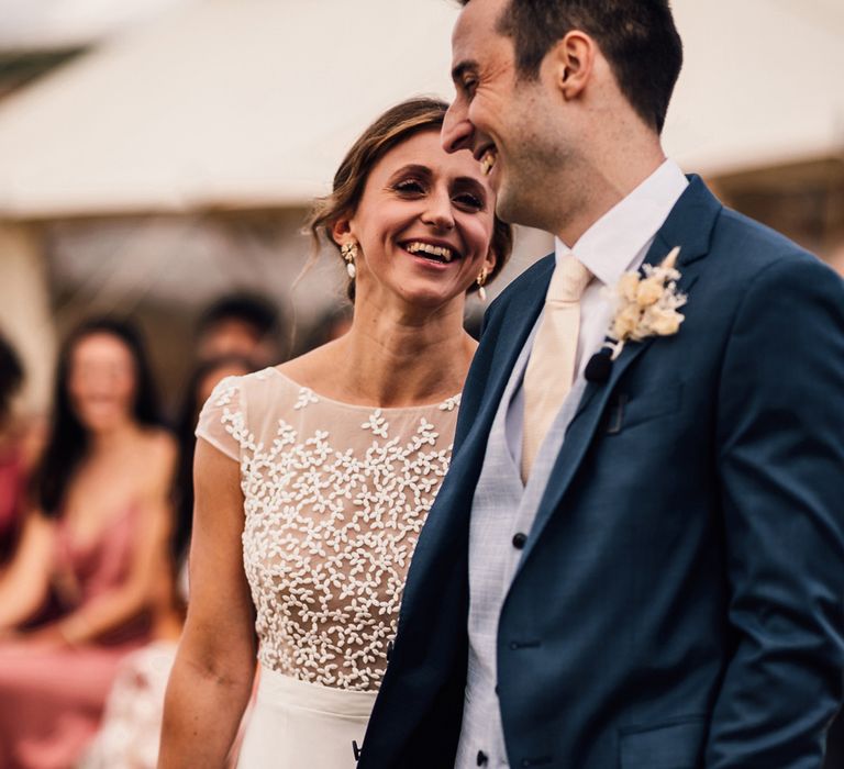 Smiling bride in white Rime Arodaky wedding dress holding dried bridal bouquet looks at laughing groom in blue three piece suit with dried floral buttonhole during outdoor wedding ceremony in Dorset