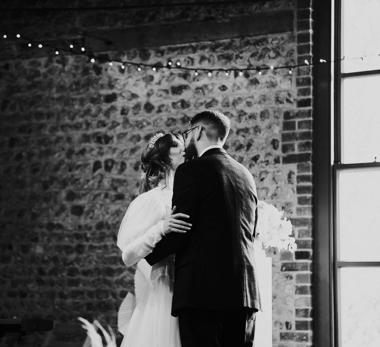 Bride & groom kiss in front of brick wall in black & white image on their wedding day