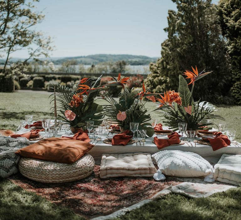 Tropical boho wedding table with low white table, floor cushions, tropical flowers and red napkins all set outside for birds of paradise wedding inspiration