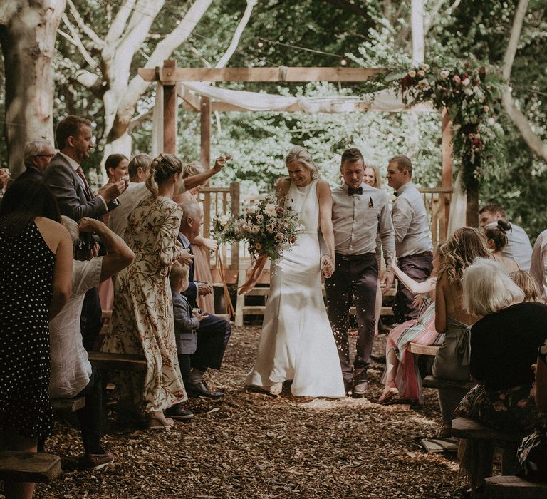 Bride & groom walk hand in hand down aisle after marrying outdoors in rustic woodland ceremony