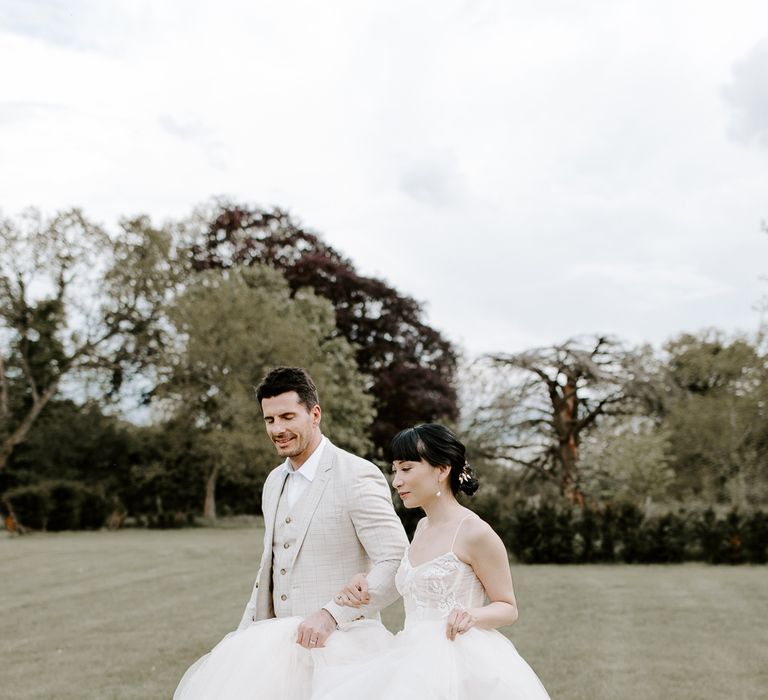 Groom in a beige check suit helping his bride hold her full skirt princess wedding dress across the field 