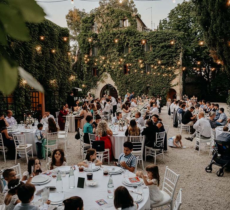 Wedding guests sit outdoors during wedding reception surrounded by villa  | Royal Studio