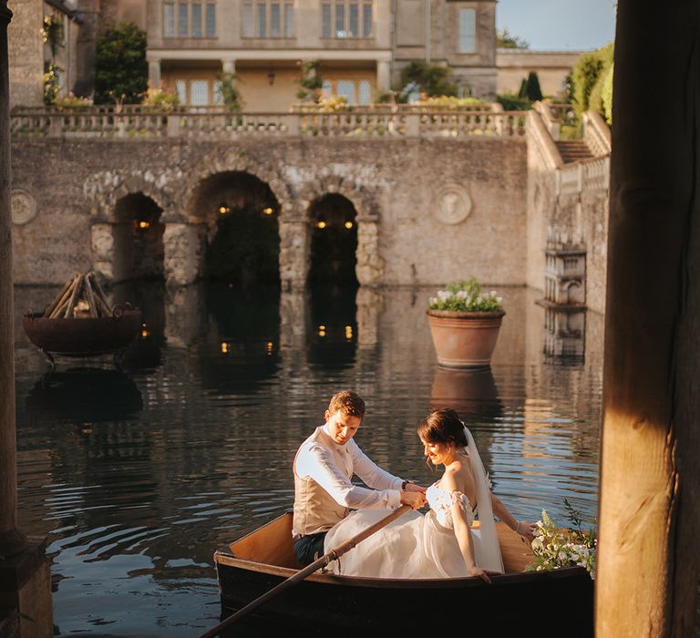 Bride & groom sit with one another in wooden boat outside Euridge Manor across the lake