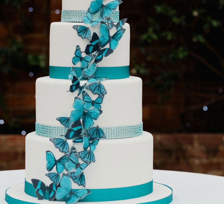 Four tier white iced wedding cake with teal blue butterfly decor and porcelain people cake topper