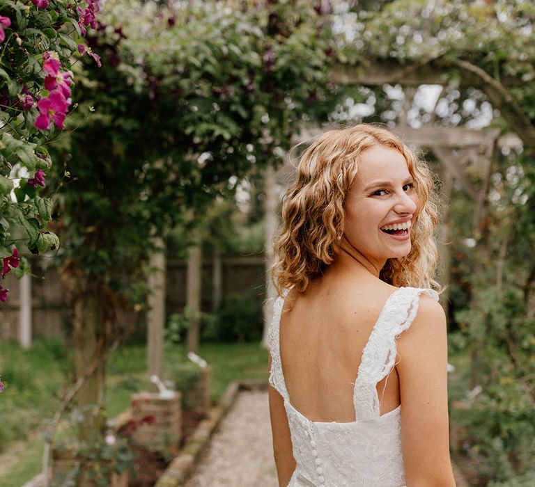 Bride looks back across her shoulder wearing her blonde hair naturally curly and lace wedding gown