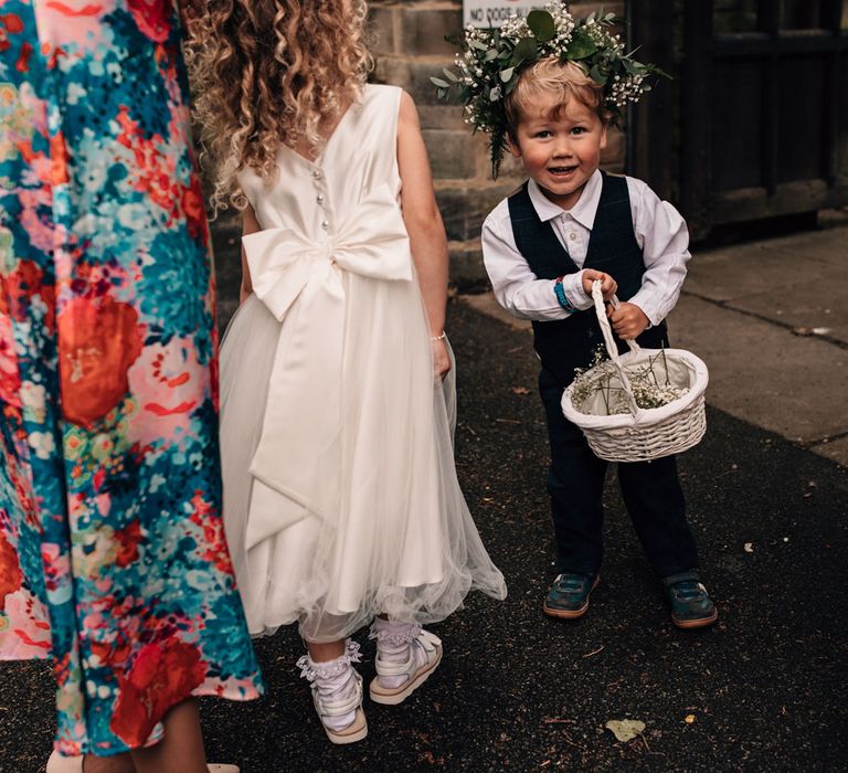 Flower boy in waistcoat and flower crown holds white basket with flower in before church wedding ceremony