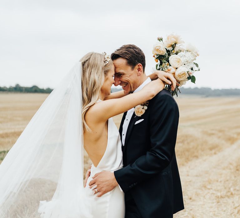 Bride in halterneck Halfpenny London wedding dress and veil holding white and peach wedding bouquet puts her arms around groom in black tie as they stand in field after wedding