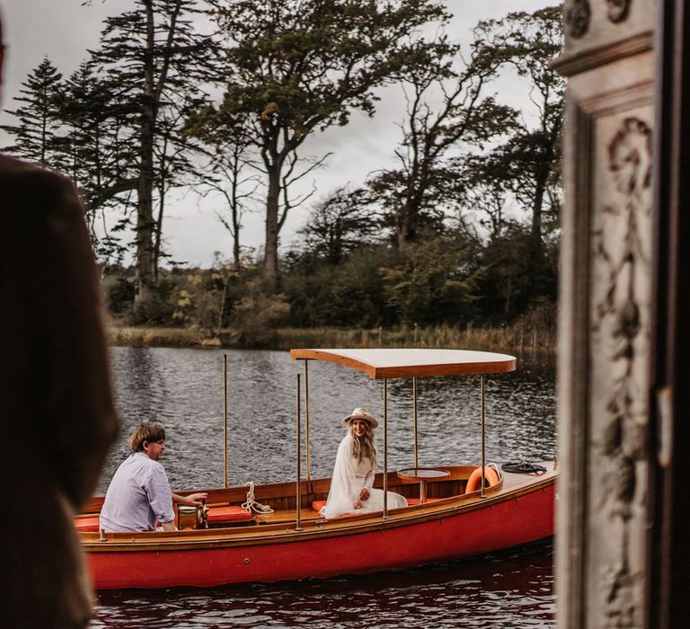 Bride in hate and tasselled bridal top sits in boat whilst groom waits on the shore of lake