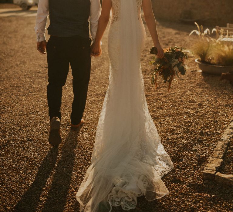 Bride in open back lace wedding dress with train and veil walks holding hands with groom in white shirt and waistcoat during golden hour