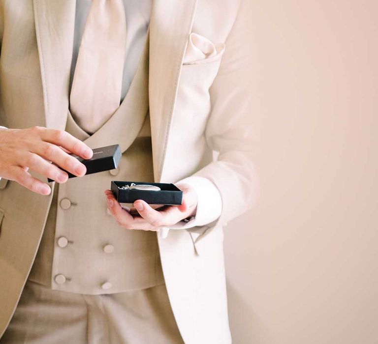 Groom wears beige wedding suit and holds pocket watch on the morning of his wedding day