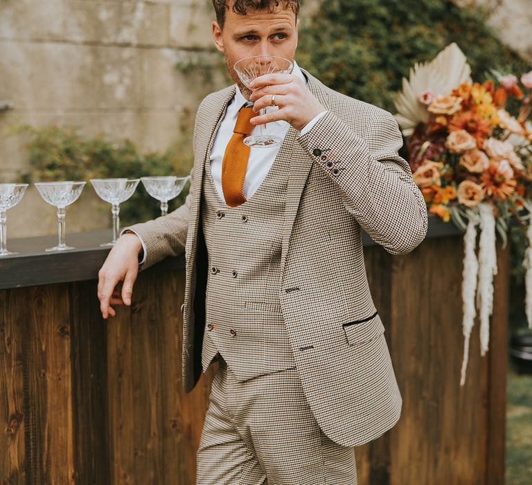 Groom in a three-piece dogstooth brown wedding suit with horseshoe waistcoat and orange tie sipping champagne at the outdoor bar 