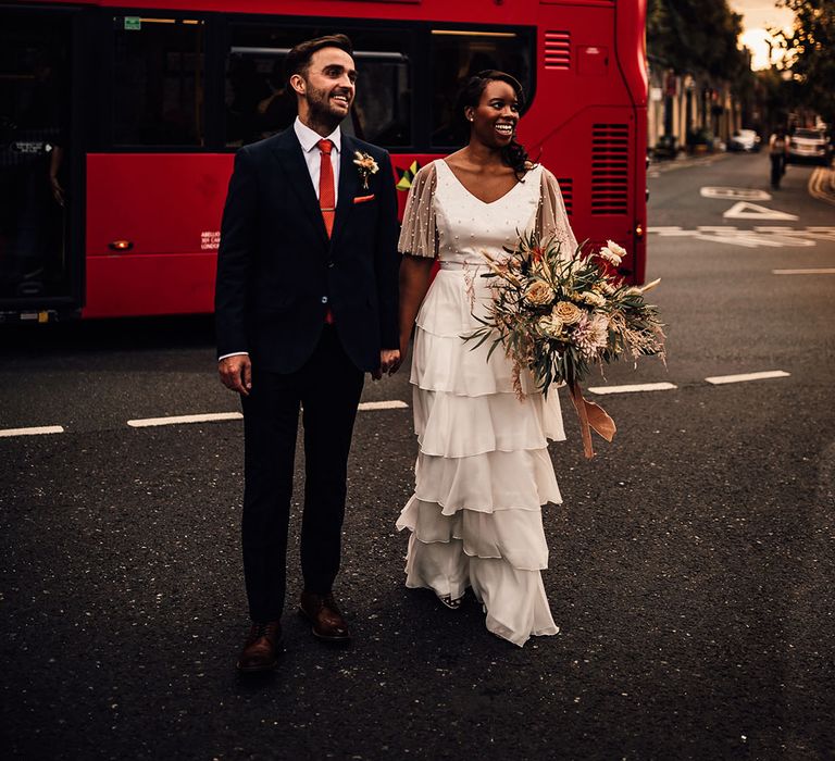 Black bride in a layered skirt and pearl top bridal separates holding hands with her groom in a navy suit in front of a red double decker London bus 