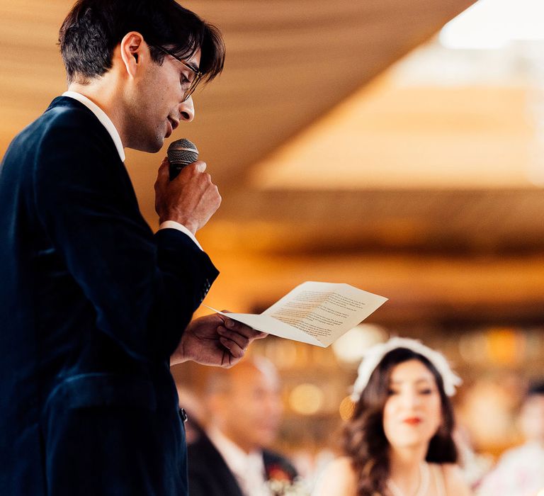 Groom in dark suit holding mic makes speech during wedding breakfast in the Cotswolds