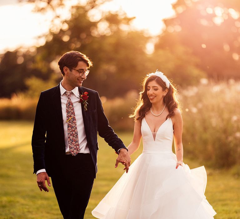 Bride in princess wedding dress with layered tulle skirt and feathered headband walks through grounds holding hands with groom in dark blue suit and floral tie during golden hour