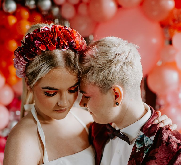 Bride with a ribbon headdress, and long eyelashes embracing her bride in a floral velvet suit jacket at retro wedding 