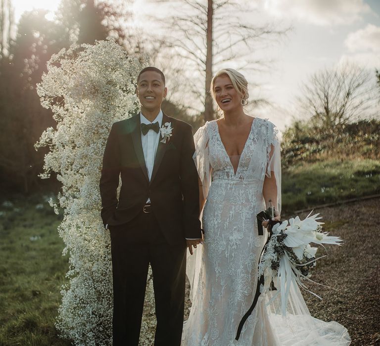 Golden hour wedding photograph of an LGBTQI+ couple holding hands at Hodsock Priory wedding in a black-tie suit and lace wedding dress