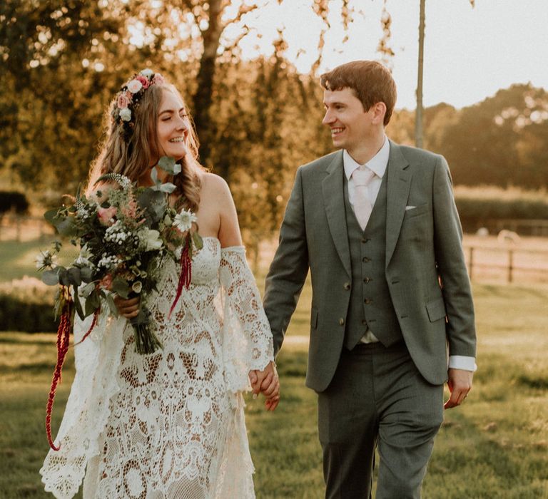 Groom in grey suit and pink tie holds hands with bride in flower crown after their woodland wedding ceremony for fantasy wedding