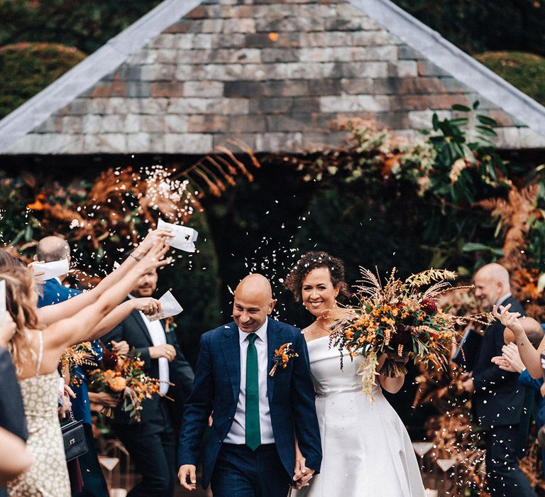 Bride in pleated off the shoulder wedding dress walks with groom in blue suit as confetti is thrown over them