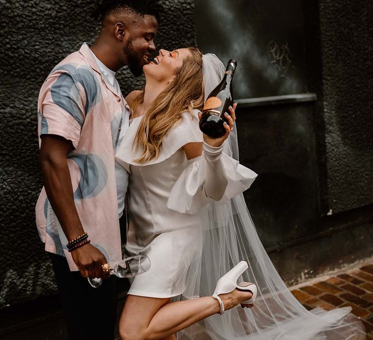 bride in a short wedding dress holding a Prosecco bottle with her groom in a patterned shirt at their city elopement 