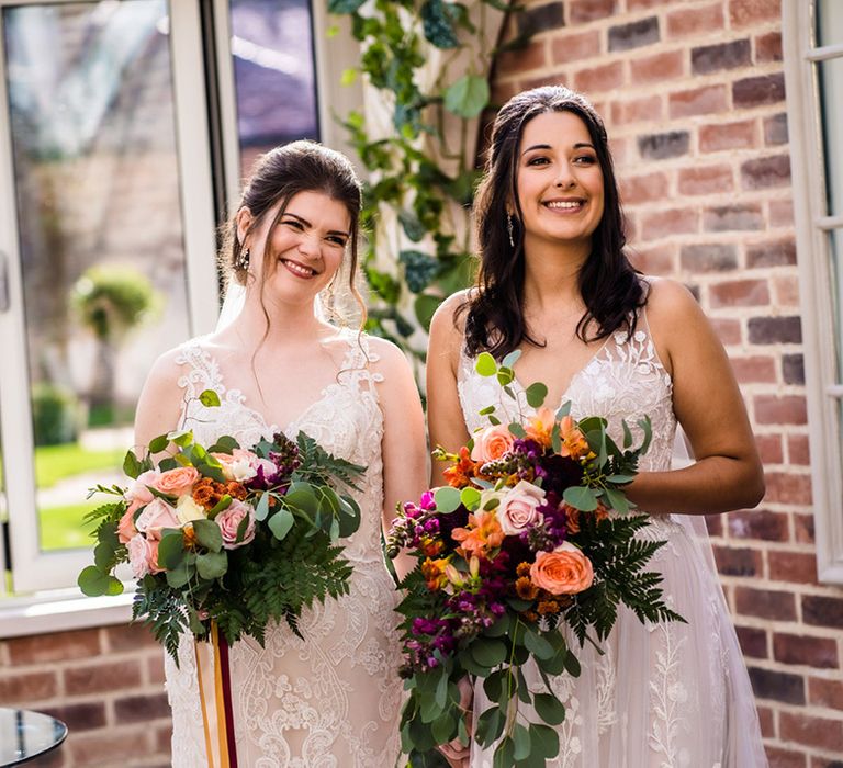 Brides stand together holding purple, pink, and orange flowers with roses and fern leaves