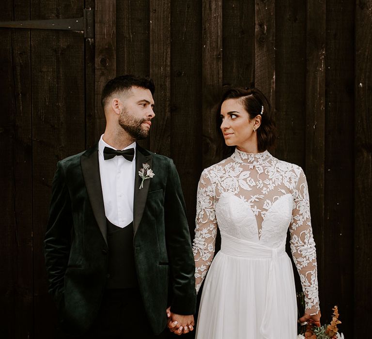 Bride with hair accessory and groom in velvet tuxedo hold hands and look at each other 