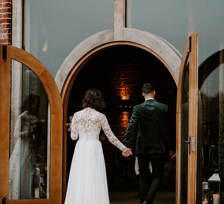 Bride in lace buttoned dress and groom in black velvet suit jacket enter their reception