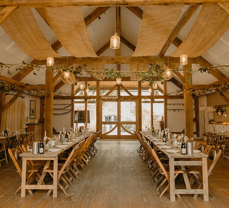 Wooden long banquet tables at rustic barn venue with decorative greenery and fairy lights 